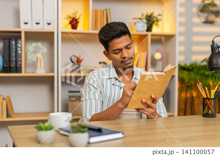 Indian man on home table reading book making notes and thinking deeply during focused study at home Indian man on home table reading book making notes and thinking deeply during focused study at home 141100057