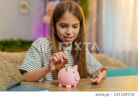Child girl kid happily saves coins into piggy bank smiling at table on home sofa planning new toy 141100158