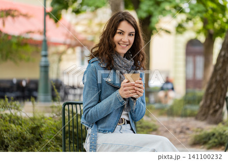 Young woman drinking takeout coffee in city street resting on park bench enjoying calm outdoor break Young woman drinking takeout coffee in city street resting on park bench enjoying calm outdoor break 141100232