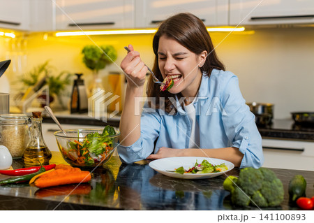 Young woman upset eating fresh salad in home kitchen struggling with healthy diet willpower concept Young woman upset eating fresh salad in home kitchen struggling with healthy diet willpower concept 141100893