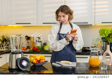 Young woman checking smartphone for food list while preparing healthy diet smoothie in home kitchen Young woman checking smartphone for food list while preparing healthy diet smoothie in home kitchen 141100937