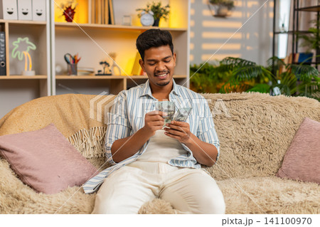 Indian man on home sofa counting cash and thinking about spending plans and future savings at home Indian man on home sofa counting cash and thinking about spending plans and future savings at home 141100970