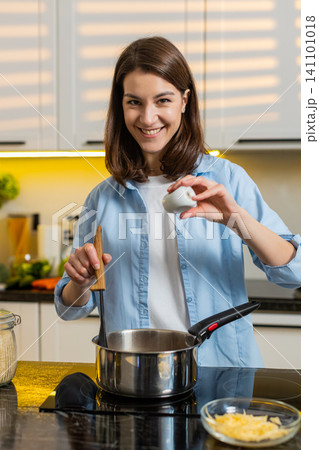 Young woman seasoning soup on stove in home kitchen smiling while cooking healthy diet recipe dinner 141101018