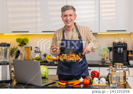 Middle-aged man frying vegetables while reading laptop screen for healthy diet meal in home kitchen Middle-aged man frying vegetables while reading laptop screen for healthy diet meal in home kitchen 141101056