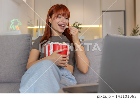 Young woman relaxes with popcorn watches funny trailer on laptop laughs smiles enjoys comedy preview Young woman relaxes with popcorn watches funny trailer on laptop laughs smiles enjoys comedy preview 141101065