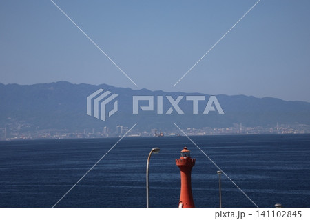 赤い灯台と青い海面の海と対岸の霞む街並みと山並みと青空の風景 赤い灯台と青い海面の海と対岸の霞む街並みと山並みと青空の風景 141102845