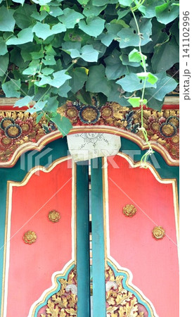 Balinese temple or house door made of red and green painted wood with gold floral carvings, a sacred mantra amulet under the arch, and vine leaves hanging from above in Bali, Indonesia. 141102996