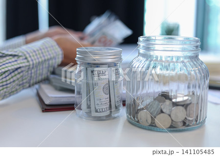 Close up glass jars filled with money and coins with businessman counting money in background. Close up glass jars filled with money and coins with businessman counting money in background. 141105485