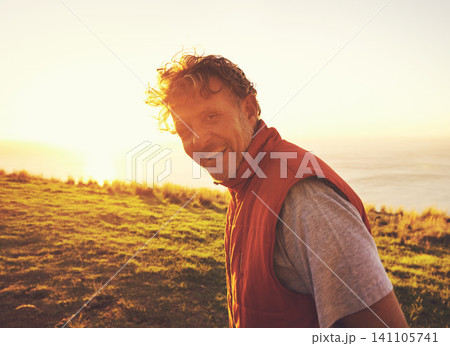 Sunset, nature and portrait of man by ocean for adventure, relaxing and enjoy weekend outdoors. Happy, smile and person with flare on hill for holiday, vacation and travel destination in evening 141105741