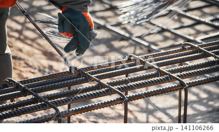 A worker is tying steel bars together to form reinforcement for a concrete foundation in a construction area. It is bright and clear outside 141106266