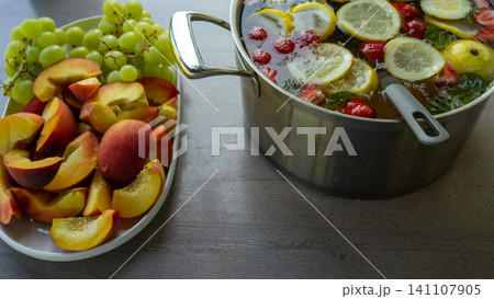 Fresh fruit assortment with sliced peaches and green grapes beside citrus berry infused water in metal pot, bright kitchen still life with healthy summer ingredients and natural colors Fresh fruit assortment with sliced peaches and green grapes beside citrus berry infused water in metal pot, bright kitchen still life with healthy summer ingredients and natural colors 141107905