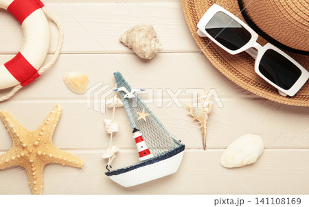Summer beach flat lay featuring straw sun hat, black sunglasses, seashells, starfish, decorative sailboat and lighthouse accent on light wooden background Summer beach flat lay featuring straw sun hat, black sunglasses, seashells, starfish, decorative sailboat and lighthouse accent on light wooden background 141108169