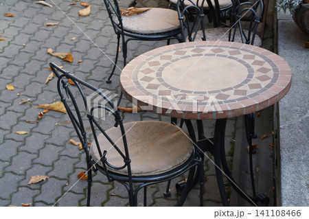 Charming Outdoor Cafe Seating Featuring a Mosaic Tiled Round Table and Decorative Wrought Iron Chairs on an Autumn Sidewalk Charming Outdoor Cafe Seating Featuring a Mosaic Tiled Round Table and Decorative Wrought Iron Chairs on an Autumn Sidewalk 141108466