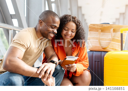 Happy young Black couple sitting together in airport corridor, checking passports Happy young Black couple sitting together in airport corridor, checking passports 141108851