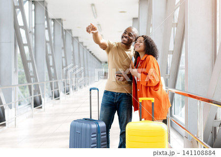 Smiling young Black couple standing with suitcases in airport, pointing toward gate 141108884