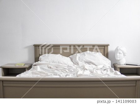 Unmade bed in a minimalist bedroom with white wall and classical bust, symbolizing rest and morning routine Unmade bed in a minimalist bedroom with white wall and classical bust, symbolizing rest and morning routine 141109083