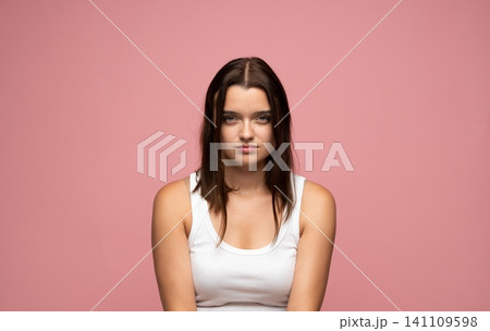 Young woman with serious expression and natural makeup looking at camera on pink background, feeling sadness Young woman with serious expression and natural makeup looking at camera on pink background, feeling sadness 141109598