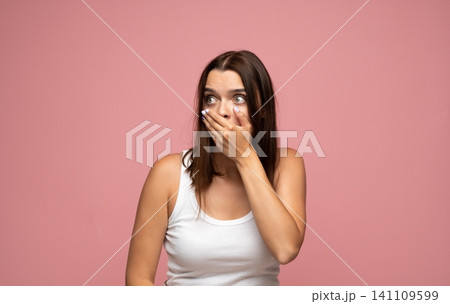 Young woman covering mouth in shock and wide-eyed surprise, expressing emotion on pink background 141109599