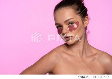 Young woman wearing pink eye patches for under eye skincare during beauty routine on pink background 141109687