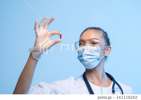 Medical professional wearing mask and safety glasses holding a red capsule, representing health, medicine, and research developing treatment 141110202