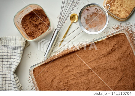 Cocoa covered tiramisu sits in rectangular glass dish beside sieve and cocoa bowl on table 141110704