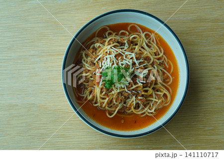 top view Pork spaghetti garnished with vegetables and a cheese slice, served in a white ceramic bowl with a black rim and placed on a wooden table 141110717