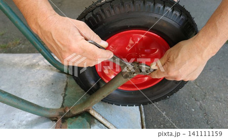 A close-up of a man's hands is shown repairing a wheelbarrow. Using an adjustable wrench, he tightens the nut on the axle of a new wheel with a bright red rim and a black, textured tire. A close-up of a man's hands is shown repairing a wheelbarrow. Using an adjustable wrench, he tightens the nut on the axle of a new wheel with a bright red rim and a black, textured tire. 141111159