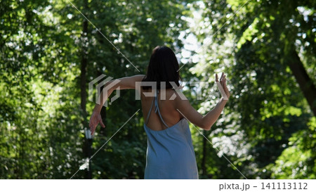A woman is dancing serenely in a park with trees and a lamp post, viewed from the rear. The scene is set in summer with a serene atmosphere, featuring green and blue ass. 141113112