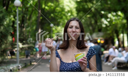 A woman enjoys ice cream in a park on a sunny summer day, with a happy expression and a front view. The scene includes a wooden spoon in a paper cup, surrounded by trees. A woman enjoys ice cream in a park on a sunny summer day, with a happy expression and a front view. The scene includes a wooden spoon in a paper cup, surrounded by trees. 141113558