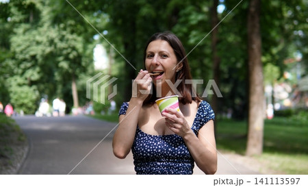 Young woman stands on shaded park path, smiling while eating ice cream from striped cup, with blurred trees, soft daylight, and calm summer atmosphere Young woman stands on shaded park path, smiling while eating ice cream from striped cup, with blurred trees, soft daylight, and calm summer atmosphere 141113597