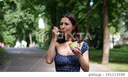 Smiling woman tastes dessert from striped cup while standing on tree lined park path, with blurred greenery, relaxed outdoor atmosphere, and bright natural daylight around her Smiling woman tastes dessert from striped cup while standing on tree lined park path, with blurred greenery, relaxed outdoor atmosphere, and bright natural daylight around her 141113599
