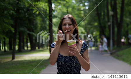 Smiling woman holding colorful ice cream cup and spoon on shaded park path, surrounded by lush green trees, enjoying relaxed summer leisure outdoors with soft natural daylight Smiling woman holding colorful ice cream cup and spoon on shaded park path, surrounded by lush green trees, enjoying relaxed summer leisure outdoors with soft natural daylight 141113611