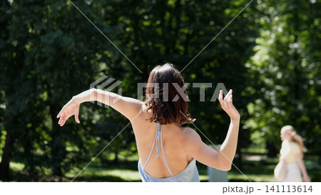 Woman seen from behind with raised arms in graceful dance pose, wearing light sleeveless top in bright sunny park with lush green trees, soft blur, relaxed summer mood Woman seen from behind with raised arms in graceful dance pose, wearing light sleeveless top in bright sunny park with lush green trees, soft blur, relaxed summer mood 141113614