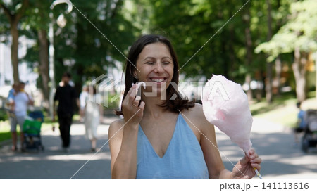 Young woman tasting pink cotton candy in bright green park, surrounded by soft focus trees and visitors, showing cheerful summer leisure, outdoor recreation and playful fair 141113616