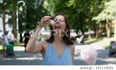 Smiling woman in blue dress tasting cotton candy in leafy park, standing on sunlit path with soft bokeh background, carefree summer moment, casual outdoor leisure and fun Smiling woman in blue dress tasting cotton candy in leafy park, standing on sunlit path with soft bokeh background, carefree summer moment, casual outdoor leisure and fun 141113638