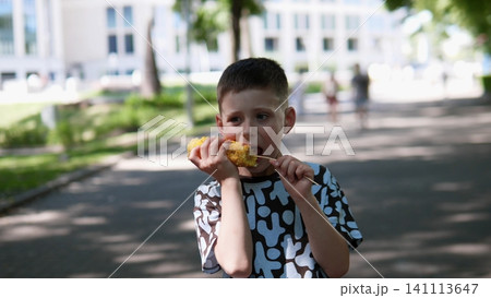 Child enjoys grilled corn outdoors on sunny summer walkway, holding cob with both hands near mouth, while soft focus background shows trees, building, and passing people Child enjoys grilled corn outdoors on sunny summer walkway, holding cob with both hands near mouth, while soft focus background shows trees, building, and passing people 141113647