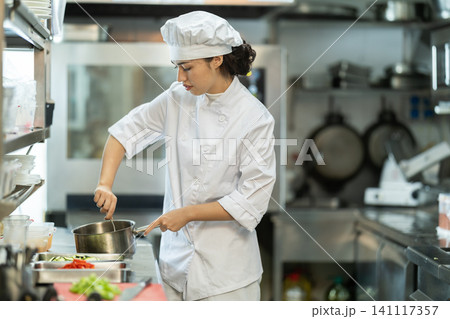 Young female cook mixing something in little saucepan Young female cook mixing something in little saucepan 141117357