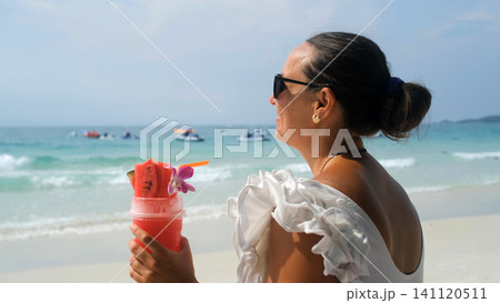 Young woman in sunglasses drinking a watermelon smoothie while relaxing in a beach chair with calm blue sea in background. Summer vacation concept Young woman in sunglasses drinking a watermelon smoothie while relaxing in a beach chair with calm blue sea in background. Summer vacation concept 141120511