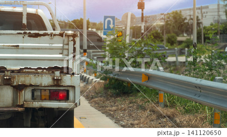 Rear view close-up of an old weathered white truck with rust stains along its tail bed. Truck is stopped on a city road lane next to a metal guardrail system. Waiting for U-Turn on the road. Rear view close-up of an old weathered white truck with rust stains along its tail bed. Truck is stopped on a city road lane next to a metal guardrail system. Waiting for U-Turn on the road. 141120650