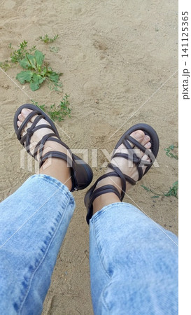 First-person top-down view of feet wearing strappy brown sandals and blue jeans, standing on sandy ground with green weeds. 141125365