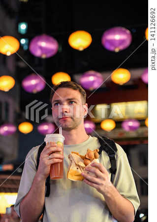 Handsome 30 years old Russian man tourist holding traditional bubble waffle and iced lemon tea at illuminated Temple Street Night Market in Hong Kong Handsome 30 years old Russian man tourist holding traditional bubble waffle and iced lemon tea at illuminated Temple Street Night Market in Hong Kong 141126692