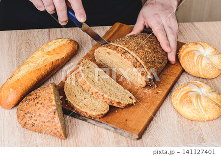 A man with a knife cuts bread on a wooden board. French baguettes. Different breeds on wooden background. 141127401