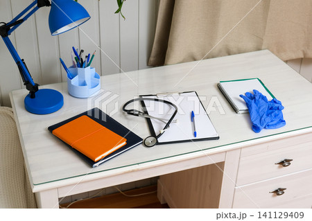 Stethoscope and blue gloves placed beside clipboard, orange notebook, pen on table. Healthcare station work space. Doctor remote workplace. Clinic telehealth workflow. Medical equipment desk Stethoscope and blue gloves placed beside clipboard, orange notebook, pen on table. Healthcare station work space. Doctor remote workplace. Clinic telehealth workflow. Medical equipment desk 141129409