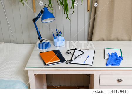 Stethoscope and blue gloves placed beside clipboard, orange notebook, pen on table. Healthcare station work space. Doctor remote workplace. Clinic telehealth workflow. Medical equipment desk Stethoscope and blue gloves placed beside clipboard, orange notebook, pen on table. Healthcare station work space. Doctor remote workplace. Clinic telehealth workflow. Medical equipment desk 141129410