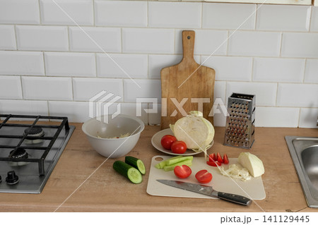 Simple home cooking. Wooden table with fresh vegetables, sliced cabbage, halved tomatoes, cucumber, cutting board knife. Preparing nutritious midday meals. Creating nourishing. Assembling hearty lunch 141129445