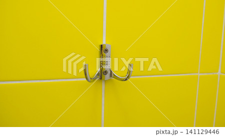 Silver metal cloth on tiled yellow wall. Strong Steel Wall Hanger. Chrome Hanger hook mounted on tile background. Mock-up. Iron clothes hook is mounted on a light wall. Ideal for hanging clothes. Silver metal cloth on tiled yellow wall. Strong Steel Wall Hanger. Chrome Hanger hook mounted on tile background. Mock-up. Iron clothes hook is mounted on a light wall. Ideal for hanging clothes. 141129446