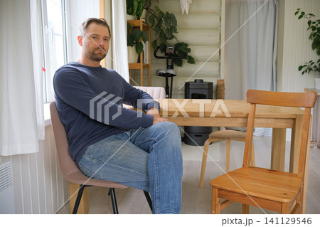 Reflective man relaxes. Middleaged male peacefully sitting inside. Indoor scene tranquil middle aged man dressed casually. Bearded 40s man seated indoors, neutral expression, wood panel background 141129546