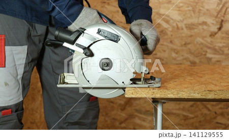 Builder saws a board with a circular saw cutting a wooden plank in a joinery. Carpenter uses portable circular saw tool to cut wood at wooden table in workshop. Worker sawing pine board. Real image 141129555