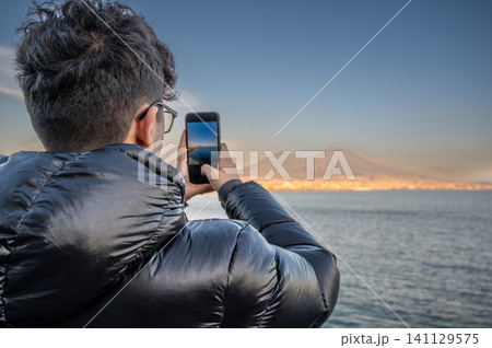 Naples, italy. December 27, 2024. Young man standing near the sea, capturing the iconic mount vesuvius and the city of naples with his mobile phone Naples, italy. December 27, 2024. Young man standing near the sea, capturing the iconic mount vesuvius and the city of naples with his mobile phone 141129575
