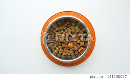 Pet food grains. Dry animal food bits in an orange metal bowl on a white studio background. Cat or dog kibble heap. Creative funny image for ad of pet products. Top view. Carnivore of domestic animals Pet food grains. Dry animal food bits in an orange metal bowl on a white studio background. Cat or dog kibble heap. Creative funny image for ad of pet products. Top view. Carnivore of domestic animals 141130927